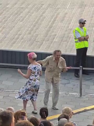 A group of people are standing on a paved outdoor area. In the foreground, several people are visible from the shoulders up, facing away from the camera. Behind them, a man with short brown hair in a blue t-shirt and denim shorts is talking to a woman with pink hair wearing a white and colorful dress. To their right, an older man in a tan button-down shirt and light-colored pants is dancing or gesturing. Further to the right, a person wearing a bright yellow safety vest and a grey baseball cap stands with their arms crossed. The background shows a large, light-colored paved surface with a dark border.