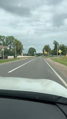 The video is filmed from the perspective of a driver looking forward through the windshield of a white car. The car is traveling on an asphalt road with a white line marking the lane. On the left side of the road, there are houses with white fences and trees. On the right side of the road, there is grass and trees, with utility poles visible. Several road signs are present, including a speed limit sign indicating 40, a school crossing sign with children figures, and a yield sign. In the distance, other cars and a few individuals can be seen. The sky is overcast with grey clouds. The overall environment appears to be a rural or suburban setting.