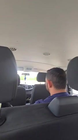 A video from inside a van shows the back of a man's head as he sits in the driver's seat. In the foreground, another person's legs are stretched out, with their feet resting on the dashboard. The person is wearing black athletic shoes and dark shorts. Outside the van, trees and greenery are visible through the windows. The video pans slightly to show the passenger in the front seat and the view outside.