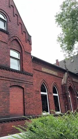 The video shows the exterior of an old brick building, possibly a school or church, with arched windows and decorative brickwork. Lush green trees and bushes surround the building, creating a natural and slightly overgrown atmosphere. The sky is overcast, suggesting a cloudy day. The camera pans slowly across the facade, highlighting the architectural details and the surrounding foliage. There are no people visible in the video.