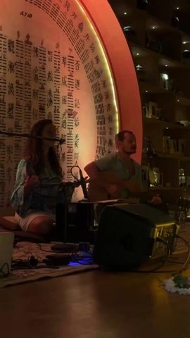 Here's a description of the video:

A man and a woman perform live music in an intimate setting. They are seated on a patterned rug on a wooden floor. The woman sings into a microphone, gesturing with her hands, while the man plays an acoustic guitar. A speaker and some small electronic devices are on the floor in front of them. 

Behind the performers is a large circular wall decoration with intricate Chinese characters, illuminated with a warm orange light. To the right, a tall shelving unit displays various objects, softly lit. A lit candle and decorative lamp sit on the floor next to the speaker.