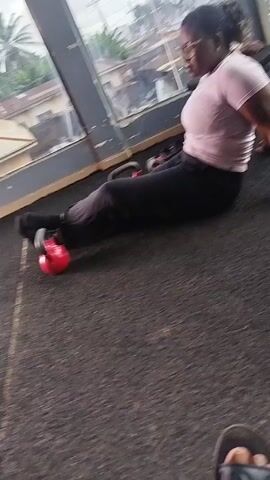 A woman is performing an exercise on the floor in what appears to be a gym. She is wearing a pink shirt and black pants, with black socks. A red kettlebell is positioned next to her feet. She is leaning back on her hands, with her legs extended and bent at the knees. The gym has a dark gray floor and a large window with a view of buildings and trees outside. The lighting suggests an overcast day.