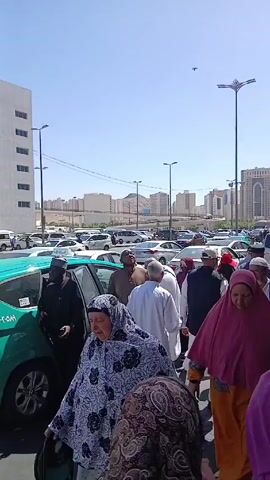 A crowded outdoor scene unfolds on a sunny day, revealing a diverse group of individuals, mostly dressed in traditional Islamic attire. Many women are wearing hijabs in various colors and patterns, while men don white robes and head coverings. They are milling around a bright teal taxi with Arabic writing on its door, which is in the process of picking up passengers. The foreground is filled with people moving in different directions, some looking directly at the camera.

In the background, a busy parking lot is packed with a variety of cars. Beyond the parking lot, the cityscape includes a mix of white buildings, with some high-rise structures, and a distant mountain. Street lights are visible along the road, adding to the overall urban setting. The sky is a clear, bright blue.