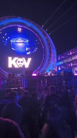 The video displays a large outdoor event with a stage and a crowd. A circular screen on the stage shows the letters "K V" in large font, with a stylized image of a man's face. The screen is illuminated with blue and purple lights. The stage itself is decorated with bright purple lights. A DJ booth is visible on the stage with several people. The crowd in front of the stage is silhouetted against the lights and appears to be watching the performance. The overall atmosphere is energetic and festive.