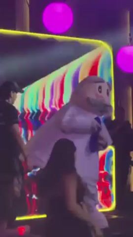 Here's a detailed description of the video:

The video shows a mascot character dressed in white with a cartoonish face, seemingly at an event or party. The mascot has a tie and is standing among people, some of whom are interacting with it. A person in a black shirt adjusts the blue element on top of the mascot's head. The background is illuminated with neon lights and large pink orbs. There is a stage-like setup with various people walking around. The scene appears to be lively and the lighting creates a vibrant atmosphere.
