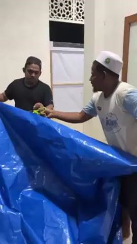 Two men hold a large blue tarp. A third man holds green scissors and cuts the tarp. The man with the scissors wears a black t-shirt and a black cap with gold embroidery. The man on the right wears a white t-shirt with a graphic and a white cap. The man on the left wears a black t-shirt. The background includes white walls, a partially visible window with an ornate design, and a partially visible white structure.