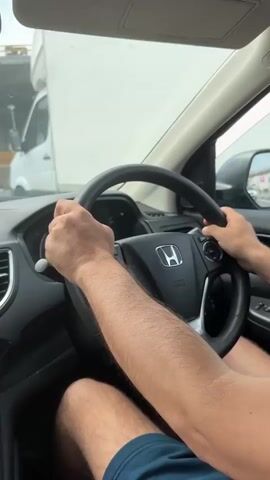 A first-person perspective video shows a person driving a Honda vehicle, presumably a CR-V, on a highway. The driver, a male, has short brown hair and is wearing blue shorts. His hands are visible on the steering wheel. Outside the car, there is a truck, and construction activity with equipment, including cranes. The dashboard has a navigation system on a cell phone that shows a route. The weather appears to be overcast.