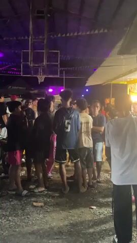 A group of people stand in a loose gathering under a partially covered outdoor space with a basketball court. The lighting shifts through shades of blue, purple, orange, and white, highlighting the crowd and casting shadows on the ground, which appears to be gravel or dirt. Some people are standing still, while others are moving slightly, engaging in conversation. A food or vendor stall is visible to the right, illuminated by a bright light. A parked motorcycle is partially visible on the left.
