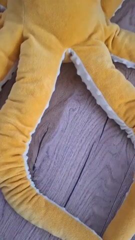 A close-up, top-down shot shows a plush yellow octopus toy with large, white and black eyes. The octopus is lying on a wooden floor, with its tentacles spread out. The toy has a textured appearance, and the inside of its tentacles is lined with white material.