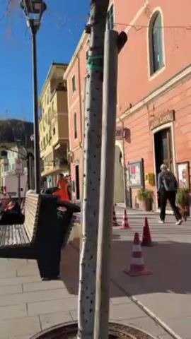 The camera focuses on a street in an Italian town. There are several workers wearing orange safety vests working on the pavement. There is a bench in the foreground, and construction equipment can be seen nearby. The buildings on the street are painted in various colors, including yellow and pink. There is a digital clock on a balcony that reads 14:53. The weather is sunny and clear. There are trees in the foreground. People walk by in the background.