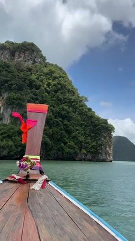 A person is on a longtail boat, cruising through turquoise waters. The boat is decorated with flowers and colorful ribbons. Towering limestone karsts covered in lush green vegetation rise dramatically from the sea. The sky is a vibrant blue, dotted with fluffy white clouds. The camera pans across the scenery, capturing the natural beauty of the tropical landscape.