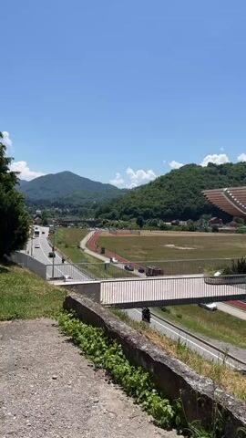 The video is a landscape shot of a modern building with a curved roof and a pedestrian walkway. The building is situated in a valley with rolling green hills in the background. A road and a running track are visible in the foreground, with a few cars and people on the track. The sky is clear and blue with a few scattered clouds. The overall impression is one of a peaceful and scenic setting.