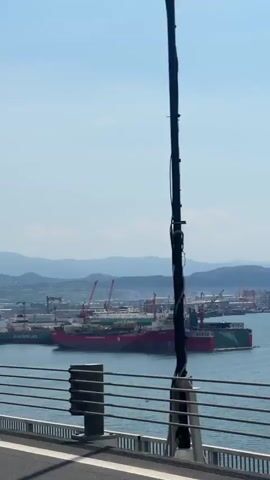 The camera moves along a bridge with views of a large port, possibly a shipyard, in the distance. Several large ships are visible in the water, and numerous cranes dot the landscape near the port. The day is bright, with a clear blue sky and a few distant mountains. Bridge infrastructure, including metal railings, poles, and transparent barriers, are in the foreground. A blue sign with a telephone icon and the words "ACIL YARDIM SOS" is attached to one of the poles. The camera captures the view as it passes, showcasing both the infrastructure of the bridge and the distant port activity.