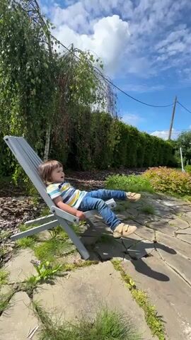 A young child lies in a gray lounge chair outdoors on a sunny day. The child is wearing a striped t-shirt and blue jeans. The chair is positioned on a stone patio, with a large tree to the left and a green hedge to the right. The sky is blue with white clouds.