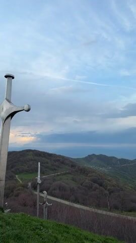 The video shows three large metallic swords planted on a hilltop overlooking a forested mountain range. The sky is partly cloudy with streaks of light peeking through the clouds. The foreground consists of green grass and bare dirt. A voice is heard in the background, but the words are unintelligible.