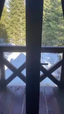 A hand opens a dark brown wooden door from inside a room, revealing a balcony view of a snow-covered landscape. The balcony has a wooden railing with an X pattern. In the distance, there's a dense forest of tall, green evergreen trees. The sky is blue and clear. Snow covers the ground below the balcony. There are lights strung along the railing.