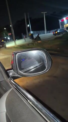 At night, a black SUV drives past a parked car, as seen from the parked car's side mirror. Streetlights illuminate the scene, revealing people standing nearby and sitting on a motorcycle. Another car is parked further down the road. The environment appears to be a street or parking area near a building.
