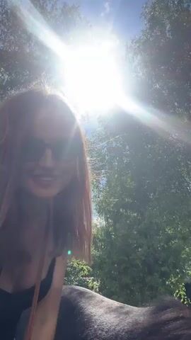 A woman in sunglasses is taking a selfie with a black horse in a park. She is smiling and sitting next to the horse. A young man is kneeling behind the horse, looking at the camera. The sun is shining brightly through the trees.