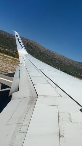 The video shows a close-up view of an airplane wing during takeoff or landing. The wing is white with the Ryanair logo visible on the wingtip. The camera is positioned to give a view of the wing, runway, and a mountainous landscape in the background. The sky is a clear blue. The video appears to be a short clip, possibly from a passenger's perspective, offering a glimpse of the flight experience. There are no people visible in the frame, and the focus is on the aircraft and its surroundings. The overall impression is one of motion and travel.