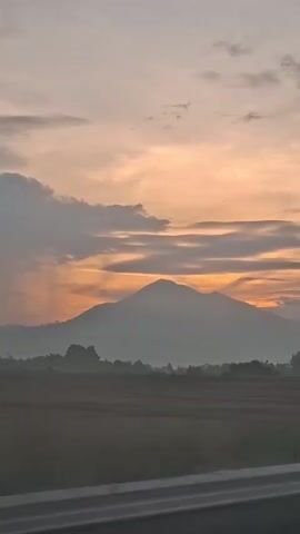 This video shows a serene landscape at sunset. In the foreground, there is a road or highway, suggested by a gray barrier. Beyond the barrier lies a field, possibly agricultural, with subtle variations in color indicating crops or soil. Dominating the mid-ground is a large, round mountain, its peak silhouetted against the sky. The mountain appears misty or hazy, adding to the atmospheric quality of the scene. The background is a dramatic sky filled with wispy clouds illuminated by the setting sun. The colors in the sky range from soft oranges and yellows near the horizon to muted grays and blues higher up. The overall impression is one of tranquility and natural beauty. There are no people or specific objects in motion, only the static elements of the landscape and the passive movement of the camera, implied by the slight angle shift and perspective.