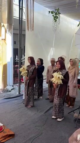 A group of people are gathered under a white tent outdoors. In the foreground, two women in matching brown outfits are holding decorative white objects. Behind them, other individuals are visible, including a man in a tan outfit and a woman with a microphone. The ground is covered with a gray mat, and a small table with bowls of colorful items is in the lower left corner.