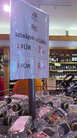 The video shows a display of small liquor bottles in a store. A sign above the bottles advertises "Fies Schnäpse und Liköre" (Fies Spirits and Liqueurs) with pricing information. The bottles appear to be a variety of types, including some that look like clear spirits and others that are brown, likely liqueurs. The camera zooms in on one of the brown bottles, which is labeled "Fies Schokolikör mit Kirsche" (Fies Chocolate Liqueur with Cherry). A hand is holding this bottle, showing it to the camera. The background suggests a retail environment with shelves of liquor bottles visible.