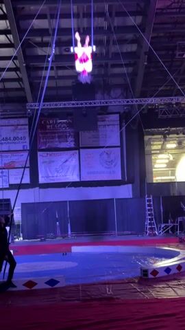 The video shows a person suspended upside down from a trapeze in what appears to be a circus tent or indoor arena. The person is wearing a sparkly, red and white outfit. The trapeze is hanging from the ceiling by two ropes, and a man stands on the floor holding a long pole, presumably for assistance or safety. The area below the trapeze is a circular stage painted with blue, white, and red patterns. Behind the stage are several large banners displaying advertisements. The arena is illuminated with blue and white stage lights. The camera zooms in on the person performing on the trapeze.