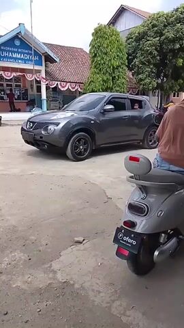 A dark grey car, possibly a Nissan Juke, is parked on the left side of the frame, facing towards the right. A person is visible inside the car, looking out the passenger-side window. In the foreground, on the right side of the frame, a light grey scooter is parked, facing towards the left. The scooter has a red accent on its rear. Behind the car, a building with blue and white siding and a red-tiled roof is visible. The building has "MADRASAH IBTIDAIYAH MUHAMMADIYAH" written on it. Several other motorcycles are parked in front of the building. Trees and other vegetation are present in the background. The ground appears to be a mix of dirt and gravel. The lighting suggests it is daytime.