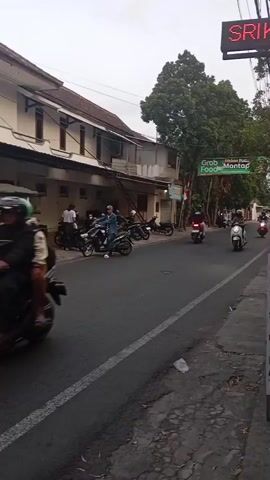 A street scene with several people riding motorcycles. On the left side of the street, there is a building with several windows. In front of the building, there are several motorcycles parked. People are standing and sitting around the motorcycles. On the right side of the street, there is a sign that says "GRAB FOOD MANTAP". There are also several motorcycles driving on the street. The sky is cloudy.