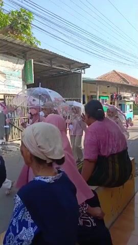 The video shows a street scene with several people. In the foreground, an older woman with a light-colored headscarf and a blue and white patterned top is sitting. To her left, another woman in a black and white headscarf is visible. In the middle ground, several women wearing pink headscarves and pink clothing are walking, some holding decorative umbrellas. One of them is holding a stick. A baby in a black shirt is being carried by one of the women. Further in the background, there are buildings with corrugated metal roofs and a sign on the wall. There are also power lines overhead. The overall impression is of an outdoor gathering or procession in a somewhat informal urban or semi-urban environment. The lighting suggests it is daytime.