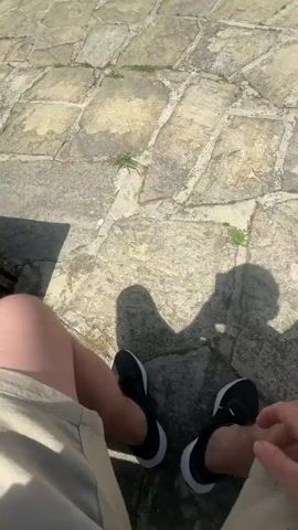 The video begins with a close-up view of a person's legs and lap, suggesting they are sitting on stone steps. The camera then pans up and to the right, revealing a courtyard paved with large, irregularly shaped stones. In the background, there is a row of buildings, trees, and a bright blue sky with fluffy white clouds. A few rose bushes with pink flowers are visible in the center of the courtyard. The overall impression is of a sunny day in a public space.