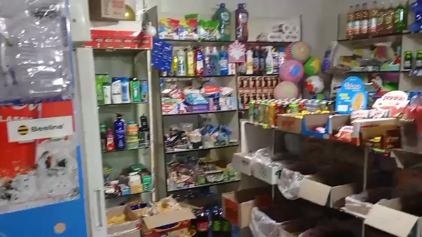 The video shows the interior of a small grocery store. The shelves are packed with various items including snacks, drinks, household goods, and candies. There is a man standing behind a counter, seemingly working. The store seems well-stocked and organized. The camera pans across the different sections of the store.