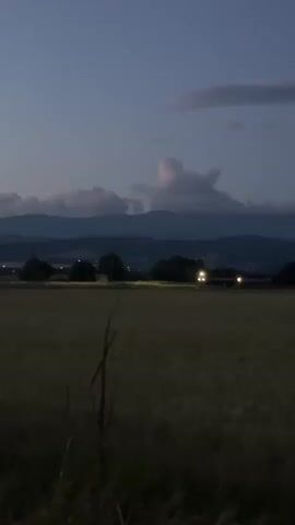 The video captures a rural landscape at dusk, likely in Galabnik, Bulgaria. The foreground is dominated by a dark field of what appears to be tall grass or crops, with a single, bare branch silhouetted against it. In the mid-ground, a line of trees and scattered buildings are visible, some illuminated by artificial lights, suggesting a small settlement or farm. Beyond this, a range of mountains recedes into the distance, their peaks softened by the fading light and possibly a light haze. The sky is a deep twilight blue, with a few wispy clouds scattered across it, one of which is catching a faint pinkish hue. The overall mood is peaceful and serene, with a sense of quiet solitude. The time of day is clearly evening, as indicated by the dim lighting and the deep blue of the sky. There is no visible text in the video.