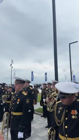 A low-angle outdoor shot captures a group of men in matching navy blue military uniforms standing in formation. They wear white caps with gold trim and dark jackets adorned with medals and gold braiding. Some hold brass instruments, suggesting they are a band. The men face forward, their expressions solemn. In the background, a cloudy sky is visible, with a formation of nine fighter jets performing aerial maneuvers, leaving smoke trails. A large white flag with a blue cross also flutters in the wind. Several tall, slender black poles stand vertically in the scene, and a few white flags with blue lettering are planted in the ground. The ground appears to be paved with light-colored tiles.
