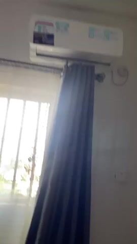The camera focuses on the upper portion of a room, highlighting an air conditioning unit mounted high on a white wall above a window. The window is covered with sheer white curtains layered behind a thicker, dark gray curtain. A curtain rod is visible above the window frame. A switch plate is noticeable on the white wall to the right of the curtain. The camera moves slightly upward, adjusting the focus on the air conditioning unit and the ceiling. The scene is well-lit, with natural light streaming in from the window.