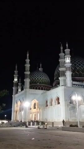 The nighttime outdoor video shows a large, brightly lit white mosque with multiple domes and minarets against a dark sky. The mosque features arched windows and intricate designs. Several streetlights illuminate the area around the mosque. A few people are visible near the mosque's entrance and on the paved ground in front of it.