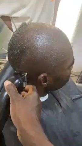 A close-up video shows a person receiving a haircut. The camera focuses on the back and side of the head of the person getting their hair cut, whose skin appears to be dark. The hair is very short and dark. A person, presumably a barber, is using an electric hair clipper to cut the hair. The barber's hand is visible, holding the black clipper with a red light indicating it's on. The person receiving the haircut is wearing a black barber cape with a white neckline. A white garment is visible behind the barber's arm. The video appears to be taken indoors, possibly in a barbershop or at home. There is a faint background noise, possibly an external sound.