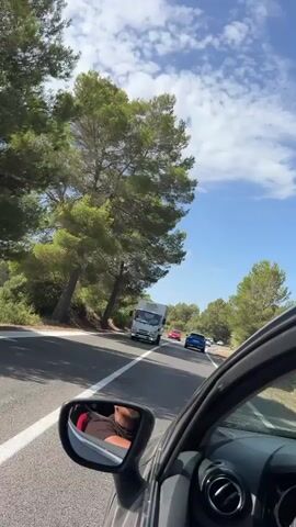 A truck is driving down a road with trees on the side. There are other cars on the road, and the sky is blue with clouds. The video is taken from inside a car, looking out the window. The driver of the car can be seen in the rearview mirror.