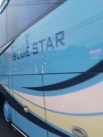 The video shows an overcast day on a paved road. The first shot is of a large blue bus. It is followed by a shot taken from a moving vehicle of a road with several parked and moving vehicles. A person riding a moped travels in the same direction as the camera. A large construction vehicle travels towards the camera. Buildings and power lines line the road. The road has a black and white striped median.