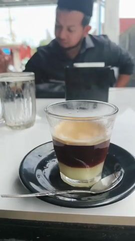 A man wearing a black cap and black shirt sits at a white table. In the foreground, a small glass of layered coffee sits on a black saucer with a spoon. To the left of the coffee is a glass of water. The man appears to be looking down at something on the table.