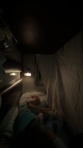 The video shows a person lying in a narrow bed in a dark room. The bed is covered with white sheets. The person is wearing a light blue shirt and is holding a pillow. There is a light on the wall above the bed. The room appears to be small and confined, possibly a train compartment or a bunk bed. The video is shaky and the lighting is dim, suggesting it was taken at night or in low light conditions.