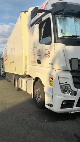 The video shows a side view of a white semi-trailer truck parked on asphalt. The truck has a large white trailer with some red lettering and logos. The truck cab is also white with some reflective sunshades visible through the windshield. The truck is positioned on the right side of the frame. The camera pans slightly to the left, revealing more of the road and a curve in the distance. Trees and vegetation are visible along the side of the road. The sky is partly cloudy with sunlight casting shadows. Another semi-trailer truck is partially visible in the distance behind the first one.