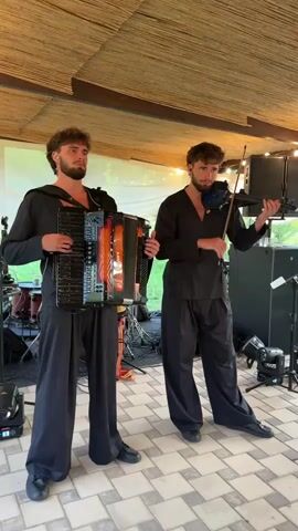 Two young men wearing black outfits and dark pants are performing music outdoors. The man on the left is playing an accordion, and the man on the right is playing a violin. They are standing on a tiled floor under a thatched roof structure. In the background, there are musical instruments, including drums, and lighting equipment. The setting appears to be a venue or outdoor performance space with greenery visible beyond.