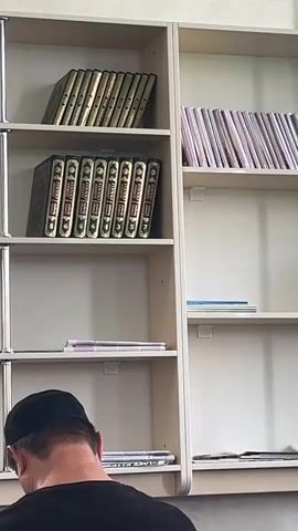 The video shows a bookshelf filled with books and a person wearing a black cap and shirt. The bookshelf is divided into several shelves. The top shelf has books with gold lettering on the spines. The second shelf from the top has a row of books with green covers and gold lettering. The third shelf is empty. The fourth shelf has a few items on it, including what appears to be a stack of CDs or DVDs. The bottom shelf has some papers and a book. A person, viewed from behind and from the neck up, is looking down at the bottom shelf of the bookshelf. The person is wearing a black t-shirt and a black cap. The environment appears to be indoors, possibly a library or a study.