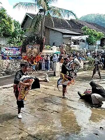 The video shows a group of performers in traditional Indonesian dance attire performing in a paved outdoor area. The pavement appears wet, possibly from a recent rain. Three men in black shirts and trousers are dancing, one performing acrobatic moves such as a handstand.  Behind them, a performer carries a large, decorative mask. A crowd of onlookers stands on a raised area, observing the performance. In the background, there's a house with a metal roof and a palm tree. The overall setting seems to be a village or rural area.