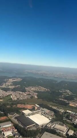 An aerial shot captures a vast landscape under a clear blue sky. The upper half of the frame is dominated by the unobstructed sky, with a faint haze visible at the distant horizon. Below, a sprawling urban and natural environment unfolds. In the far distance, a large body of water, likely a reservoir or lake, winds through hills dotted with scattered residential developments. Closer to the foreground, a patchwork of dense green forests and numerous residential areas with predominantly light-colored roofs extends across the terrain. A major road or highway, marked by dark asphalt, cuts horizontally through the mid-ground. In the immediate foreground, a cluster of large industrial or commercial buildings with flat, multi-colored roofs (white, grey, black) is prominent, alongside areas of reddish-brown earth and more residential structures. The camera maintains a stable, high-altitude perspective throughout the brief video.
