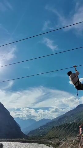 A person is zip-lining across a wide, rocky river valley. The person is wearing a white shirt, dark pants, and a helmet. They are harnessed to a wire that extends across the valley. The sun is shining brightly in the sky, and there are clouds in the distance. Mountains rise up on either side of the valley. The river below is fast-moving and appears to be shallow. The overall scene is one of adventure and excitement.