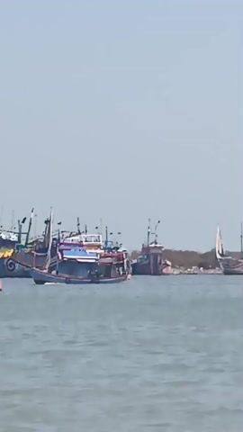 The video captures a sunny day at a harbor or coastline. The water is calm, reflecting the light blue sky. Several fishing boats are docked or moving in the water. In the foreground, a blue and red fishing boat is prominent, with a tall mast and sails. In the background, a line of larger boats, mostly blue, are moored along the shore, with some structures and trees visible on land. The overall scene depicts a typical day in a fishing community.