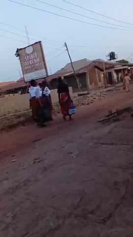 The video shows three women walking away from the camera on a dirt road in a residential area. To the left is a sign for "Spirit Afresh Ministry Intl". The women are carrying bags. A man walks away from the camera in the distance. A motorbike drives past on the right. Buildings are visible in the background. Overhead power lines stretch across the scene. The sky is overcast.