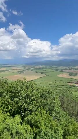 The video showcases a scenic view of a rural landscape from an elevated vantage point. Lush green fields and forests spread out under a bright blue sky dotted with fluffy white clouds. A small village with red-tiled roofs is visible in the distance, nestled amidst the greenery. The camera pans slowly, revealing the vastness of the landscape, with rolling hills and mountains forming the horizon. The foreground is dominated by dense green trees and shrubbery, suggesting the location is a hilltop or mountain peak. The sun is shining brightly, illuminating the scene and creating a sense of tranquility and openness.